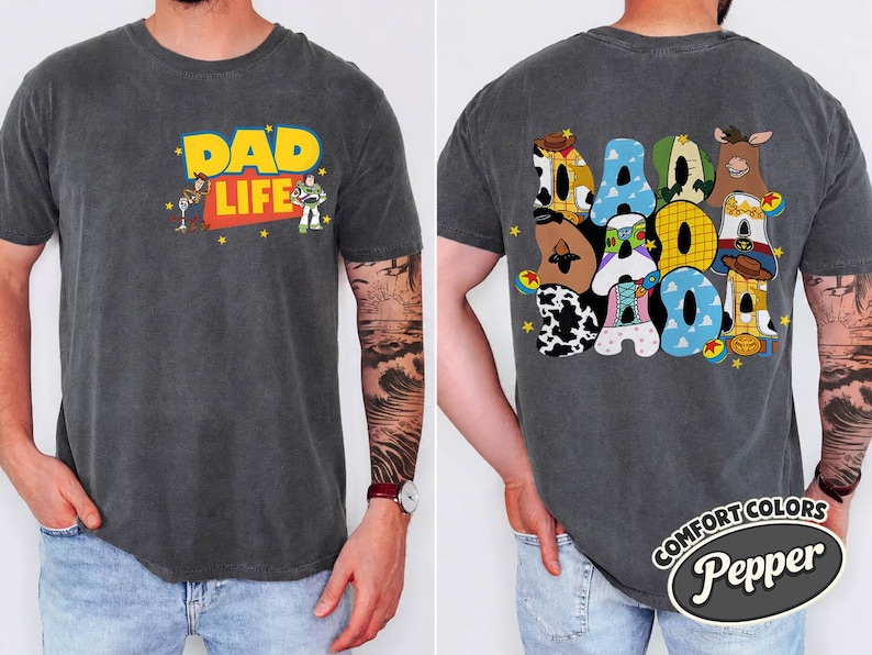 May include: Dark gray t-shirt with "DAD LIFE" in yellow and red, featuring cartoon characters. The back of the shirt displays "DAD" in colorful, character-themed letters. The shirt is labeled "Comfort Colors Pepper".