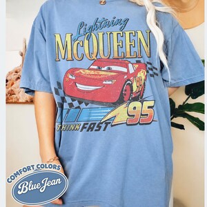 K&ouml;nnte beinhalten: Blaues T-Shirt mit kurzen &Auml;rmeln mit Lightning McQueen aus dem Film Cars. Die Grafik enth&auml;lt den Text "Lightning McQueen" &uuml;ber einem roten Rennwagen mit der Nummer 95. Der Satz "Think Fast" steht darunter.