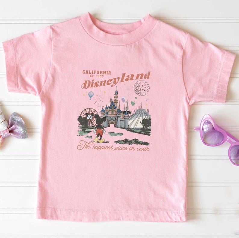 May include: Pink t-shirt featuring a Disneyland graphic. The graphic includes the text "California Disneyland Est. 1955" and an illustration of Mickey Mouse, a castle, and other park attractions. The text "The happiest place on earth" is also included.
