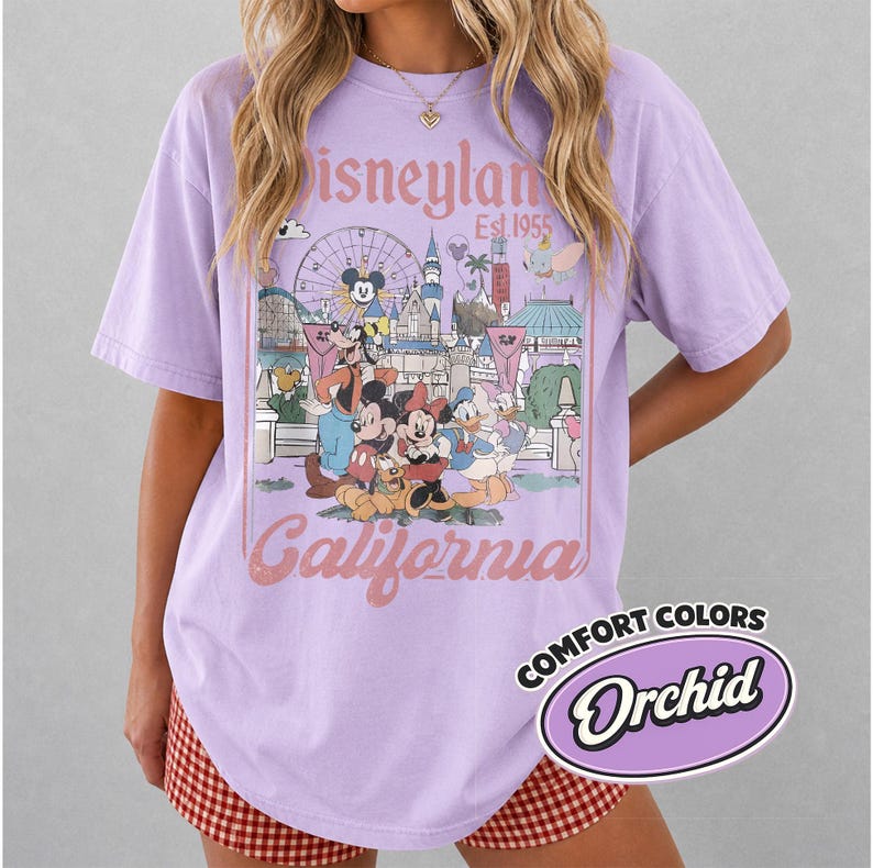 Może przedstawiać: Liliowa koszulka z grafiką Disneyland California, z Myszka Miki i przyjaci&oacute;łmi. Projekt zawiera diabelski młyn, zamek i napis "Est 1955". Koszulka ma etykietę Comfort Colors Orchid.