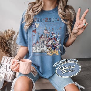 Puede incluir: Una camiseta azul con un gr&aacute;fico que celebra el 250 aniversario de EE. UU. El dise&ntilde;o presenta un castillo, personajes de dibujos animados y el texto "USA 250th Anniversary 1776-2026". La camiseta est&aacute; etiquetada como "Comfort Colors Washed Denim".