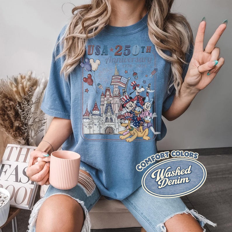 Puede incluir: Una camiseta azul con un gr&aacute;fico que celebra el 250 aniversario de EE. UU. El dise&ntilde;o presenta un castillo, personajes de dibujos animados y el texto "USA 250th Anniversary 1776-2026". La camiseta est&aacute; etiquetada como "Comfort Colors Washed Denim".