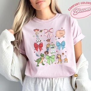 May include: A light pink crew neck t-shirt featuring a graphic print of various cartoon characters and colorful bows. The design includes a cowgirl, a cowboy, a space ranger, a green dinosaur, a pig, and a spring-bodied dog, interspersed with small stars.