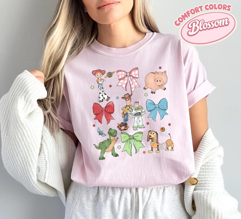 May include: A light pink crew neck t-shirt featuring a graphic print of various cartoon characters and colorful bows. The design includes a cowgirl, a cowboy, a space ranger, a green dinosaur, a pig, and a spring-bodied dog, interspersed with small stars.