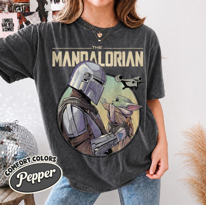 Puede incluir: Una camiseta de manga corta gris carb&oacute;n con un estampado gr&aacute;fico de The Mandalorian y Grogu dentro de un marco circular. El texto THE MANDALORIAN est&aacute; impreso en letras beige grandes sobre la ilustraci&oacute;n, que tambi&eacute;n incluye una nave espacial.