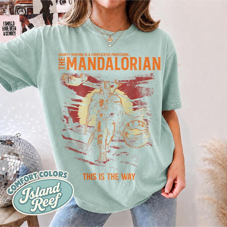 May include: A light green t-shirt featuring a graphic print of The Mandalorian. The text reads "Bounty hunting is a complicated profession... The Mandalorian" and "This is the way" in orange. The design includes a yellow sun and a small creature.