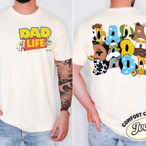May include: Ivory-colored t-shirt with the words "DAD LIFE" in yellow and blue, featuring cartoon characters. The back of the shirt displays the word "DAD" in colorful, character-themed letters. The shirt is a Comfort Colors brand.