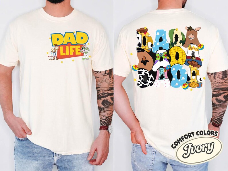 May include: Ivory-colored t-shirt with the words "DAD LIFE" in yellow and blue, featuring cartoon characters. The back of the shirt displays the word "DAD" in colorful, character-themed letters. The shirt is a Comfort Colors brand.