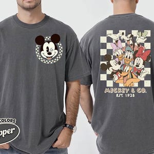 May include: Dark grey t-shirt featuring Mickey Mouse winking in a checkered circle on the front. The back displays a group of Disney characters on a checkered background, with "Mickey & Co. Est. 1928" printed below. The shirt is labeled "Comfort Colors Pepper".