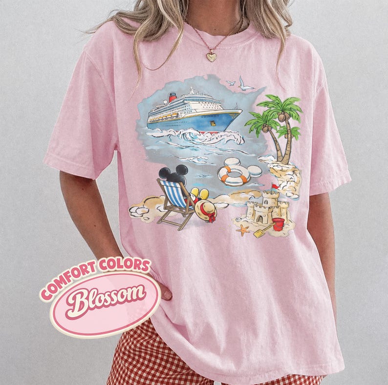Op de afbeelding: Zachtroze T-shirt met een strandsc&egrave;ne-afbeelding. De afbeelding toont een cruiseschip, palmbomen, een zandkasteel en Mickey Mouse-oren. De tekst "COMFORT COLORS Blossom" staat linksonder.