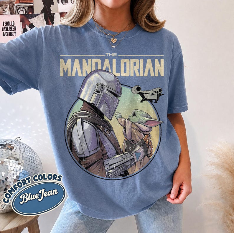 Puede incluir: Una camiseta de color azul vaquero con un estampado gr&aacute;fico de The Mandalorian y Grogu dentro de un marco circular. El texto THE MANDALORIAN est&aacute; impreso en una fuente de estilo vintage sobre los personajes, con una peque&ntilde;a silueta de nave.