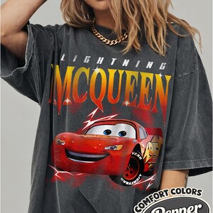 May include: Dark gray Lightning McQueen t-shirt featuring a red cartoon race car graphic. The shirt has the text "LIGHTNING MCQUEEN" in yellow and orange, and the words "COMFORT COLORS Pepper" in a white oval.