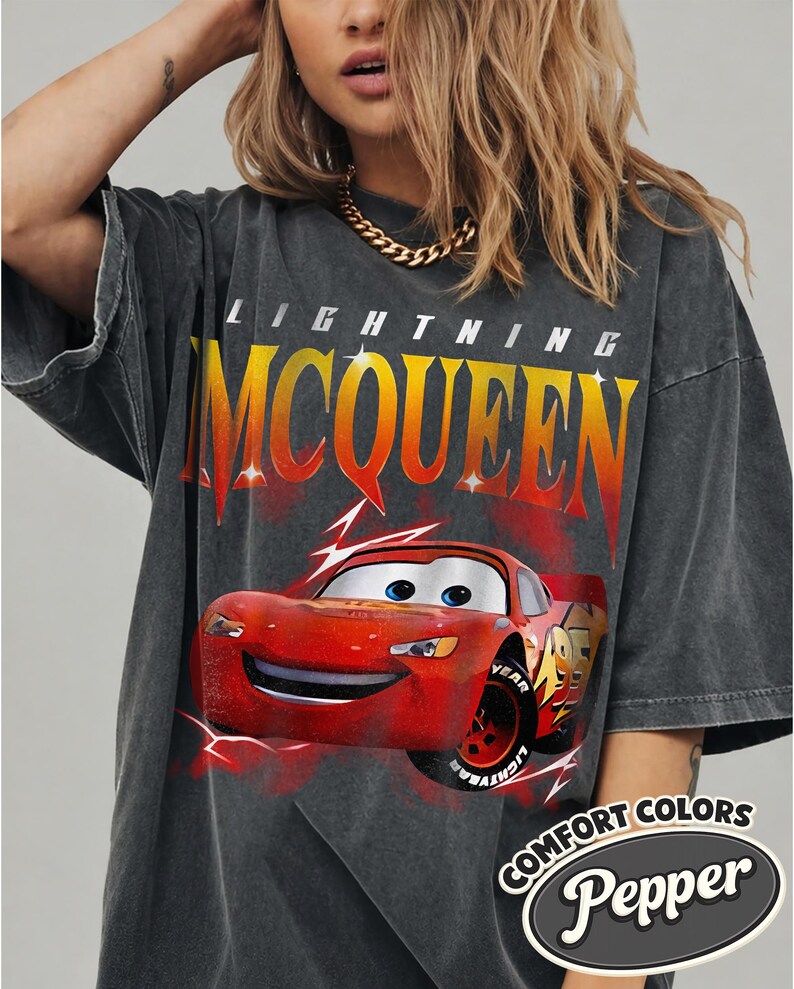 May include: Dark gray Lightning McQueen t-shirt featuring a red cartoon race car graphic. The shirt has the text "LIGHTNING MCQUEEN" in yellow and orange, and the words "COMFORT COLORS Pepper" in a white oval.