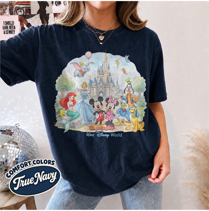 Op de afbeelding: Een donkerblauw t-shirt met korte mouwen en een kleurrijke grafische print van diverse tekenfilmfiguren voor een kasteel. De tekst Walt Disney World staat onder de figuren gedrukt. Het shirt heeft een ronde hals en een relaxte pasvorm.
