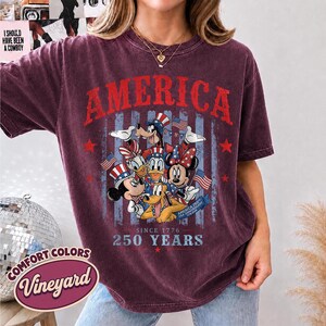 May include: A maroon short-sleeve t-shirt featuring a graphic of cartoon characters wearing patriotic hats. The text reads AMERICA, SINCE 1776, 250 YEARS, and AMERICA 250TH USA ANNIVERSARY 1776-2026. The design includes American flags and stars.