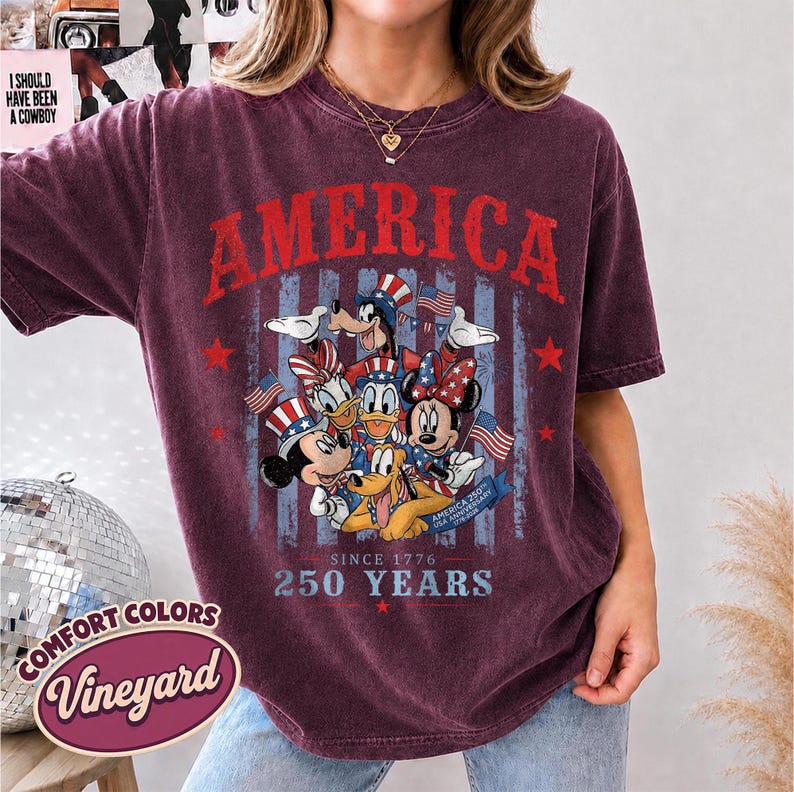 May include: A maroon short-sleeve t-shirt featuring a graphic of cartoon characters wearing patriotic hats. The text reads AMERICA, SINCE 1776, 250 YEARS, and AMERICA 250TH USA ANNIVERSARY 1776-2026. The design includes American flags and stars.