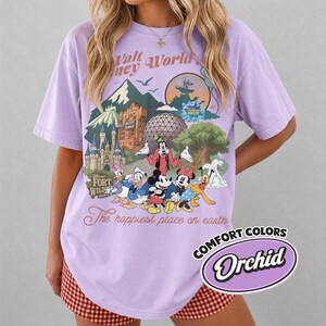 Pu&ograve; includere: Una maglietta lilla con una grafica colorata di personaggi e monumenti Disney. Il design include Topolino, Paperino e Pippo, con il testo "Walt Disney World" e "The happiest place on earth."