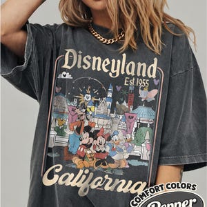 Może przedstawiać: Ciemnoszara koszulka z grafiką Disneyland z Myszką Miki, Myszka Minnie, Kaczorem Donaldem i Goofym. Koszulka ma złote napisy "Disneyland California Est. 1955". Koszulka jest oznaczona jako "Comfort Colors Pepper".