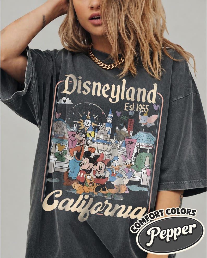 Może przedstawiać: Ciemnoszara koszulka z grafiką Disneyland z Myszką Miki, Myszka Minnie, Kaczorem Donaldem i Goofym. Koszulka ma złote napisy "Disneyland California Est. 1955". Koszulka jest oznaczona jako "Comfort Colors Pepper".