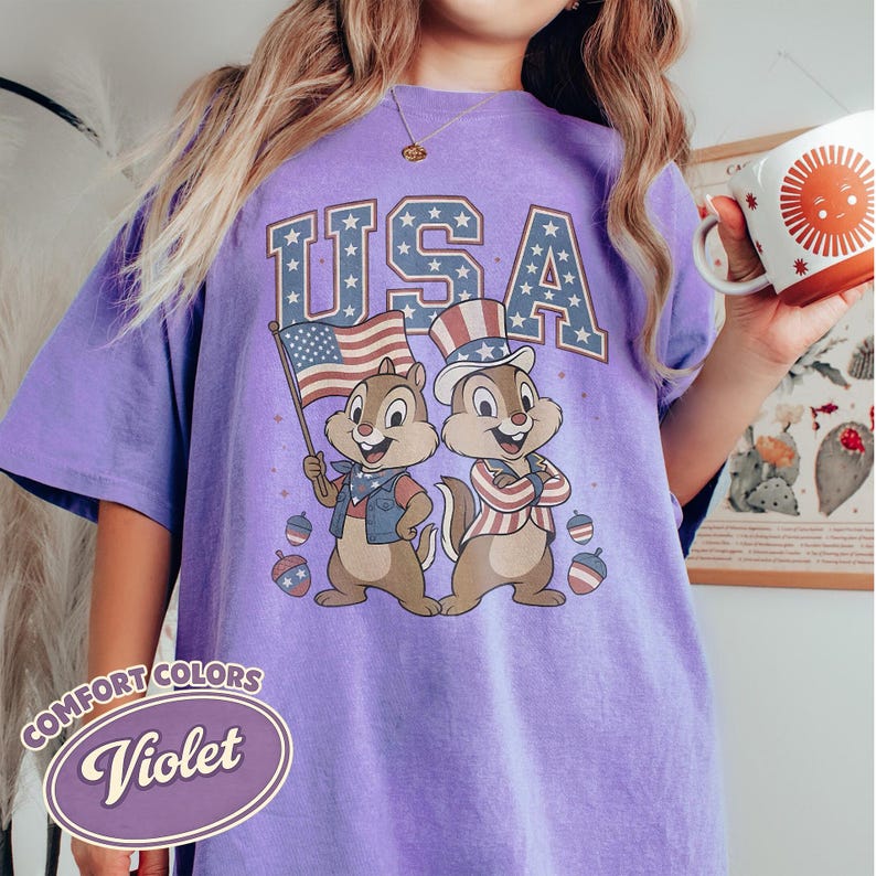 May include: Violet Comfort Colors t-shirt featuring a graphic of two cartoon chipmunks dressed in patriotic attire, with the word "USA" in blue and white stars. One chipmunk holds an American flag. The shirt has a vintage, distressed look.