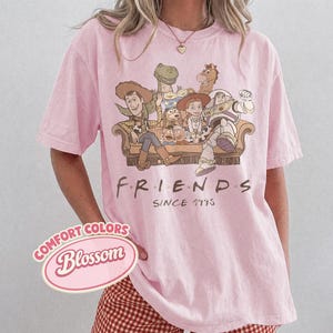 Puede incluir: Camiseta rosa claro con un dise&ntilde;o de personajes de Toy Story sentados en un sof&aacute;. La palabra "FRIENDS" est&aacute; impresa debajo del dise&ntilde;o, con "SINCE 1995" debajo. La camiseta tiene mangas cortas.