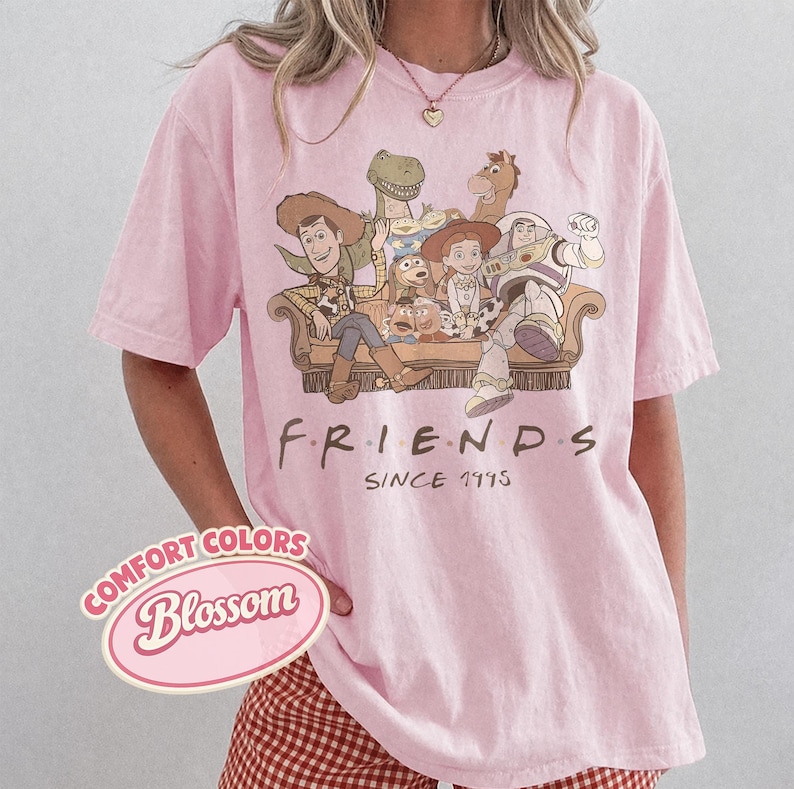 Puede incluir: Camiseta rosa claro con un dise&ntilde;o de personajes de Toy Story sentados en un sof&aacute;. La palabra "FRIENDS" est&aacute; impresa debajo del dise&ntilde;o, con "SINCE 1995" debajo. La camiseta tiene mangas cortas.