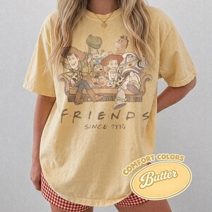 Puede incluir: Camiseta amarilla clara con un dise&ntilde;o de los personajes de Toy Story en un dise&ntilde;o tem&aacute;tico de Friends. La camiseta tiene el texto "FRIENDS SINCE 1995" y una etiqueta "Comfort Colors Butter".