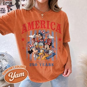 May include: An orange short-sleeve t-shirt featuring a graphic print of cartoon characters wearing patriotic hats. The text reads AMERICA, SINCE 1776, 250 YEARS, and AMERICA 250TH USA 1776-2026. The design includes American flags and red stars.