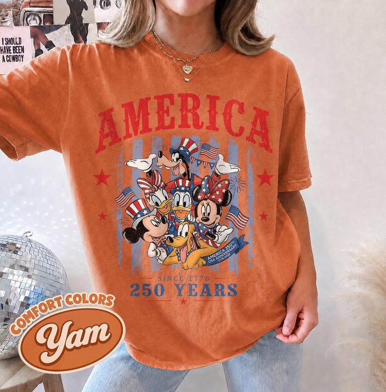 May include: An orange short-sleeve t-shirt featuring a graphic print of cartoon characters wearing patriotic hats. The text reads AMERICA, SINCE 1776, 250 YEARS, and AMERICA 250TH USA 1776-2026. The design includes American flags and red stars.