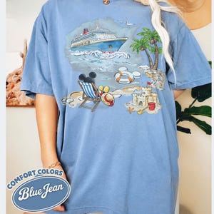 Op de afbeelding: Blauw T-shirt met een strandsc&egrave;ne-afbeelding. De afbeelding toont een cruiseschip, palmbomen, een zandkasteel en Mickey Mouse. Het shirt heeft de woorden "Comfort Colors Blue Jean" in een cirkelvormig ontwerp.