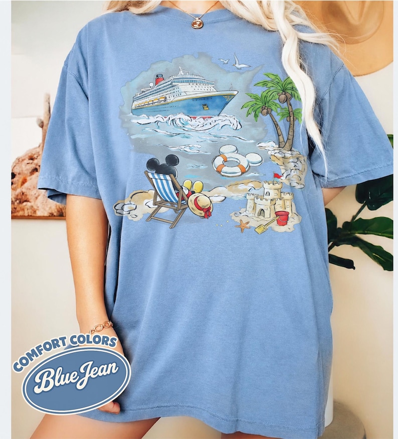 Op de afbeelding: Blauw T-shirt met een strandsc&egrave;ne-afbeelding. De afbeelding toont een cruiseschip, palmbomen, een zandkasteel en Mickey Mouse. Het shirt heeft de woorden "Comfort Colors Blue Jean" in een cirkelvormig ontwerp.