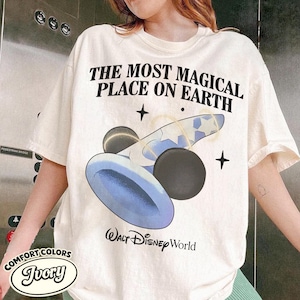 K&ouml;nnte beinhalten: Elfenbeinfarbenes T-Shirt mit dem Text "THE MOST MAGICAL PLACE ON EARTH" und einer blauen und schwarzen Grafik. Darunter steht "WALT Disney World". Das Shirt ist von der Marke Comfort Colors.