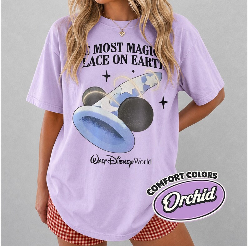 K&ouml;nnte beinhalten: Ein lilafarbenes T-Shirt mit dem Text "THE MOST MAGIC PLACE ON EARTH" und einer Grafik eines blauen und schwarzen Zaubererhuts mit Mickey Mouse Ohren. Das Shirt hat auch das "Walt Disney World" Logo und ein "Comfort Colors Orchid" Label.