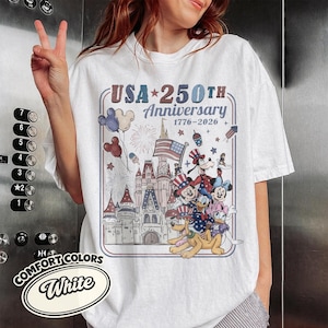 Puede incluir: Camiseta blanca con un dise&ntilde;o que celebra el 250 aniversario de EE. UU. El dise&ntilde;o presenta un castillo, Mickey Mouse, Donald Duck, Goofy y Pluto con atuendos patri&oacute;ticos, junto con fuegos artificiales y la bandera estadounidense.