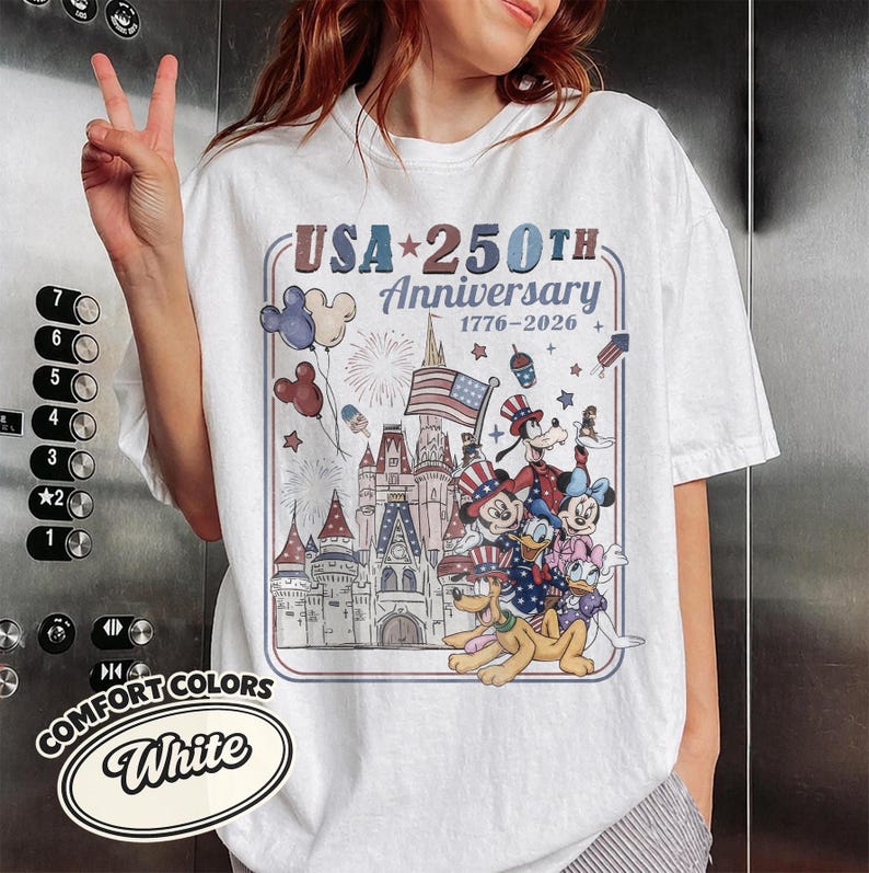 Puede incluir: Camiseta blanca con un dise&ntilde;o que celebra el 250 aniversario de EE. UU. El dise&ntilde;o presenta un castillo, Mickey Mouse, Donald Duck, Goofy y Pluto con atuendos patri&oacute;ticos, junto con fuegos artificiales y la bandera estadounidense.
