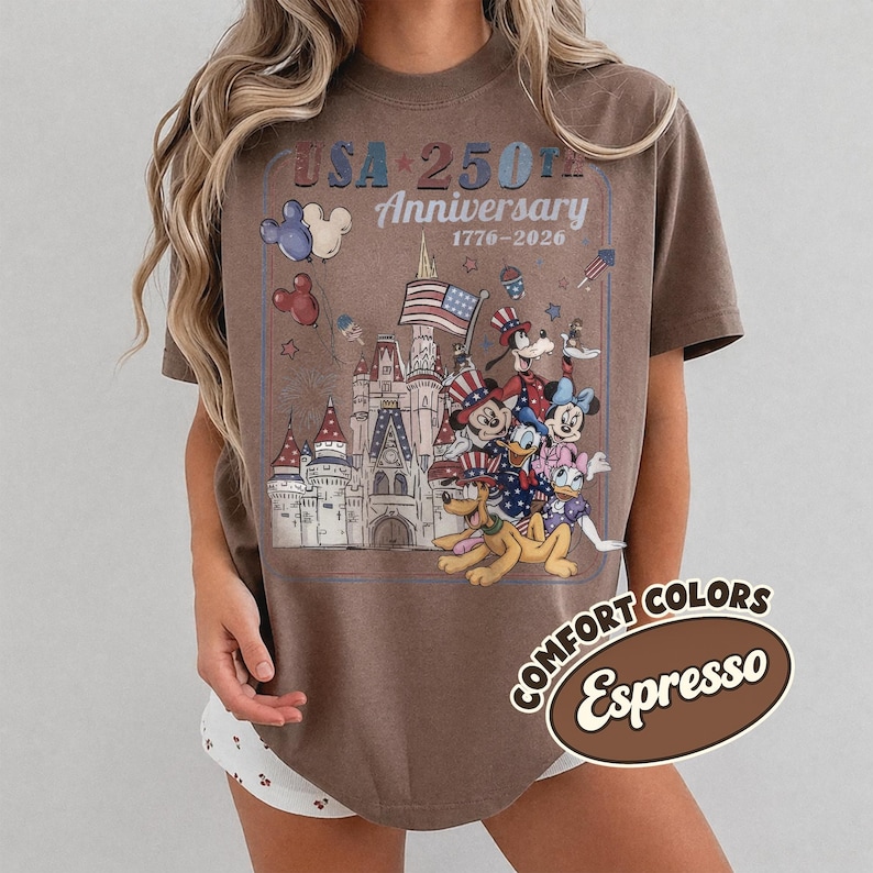 Puede incluir: Camiseta color espresso con un gr&aacute;fico que celebra el 250 aniversario de EE. UU. El dise&ntilde;o presenta un castillo, personajes de dibujos animados, la bandera estadounidense y el texto "USA 250th Anniversary 1776-2026". La camiseta es de la colecci&oacute;n Comfort Colors.