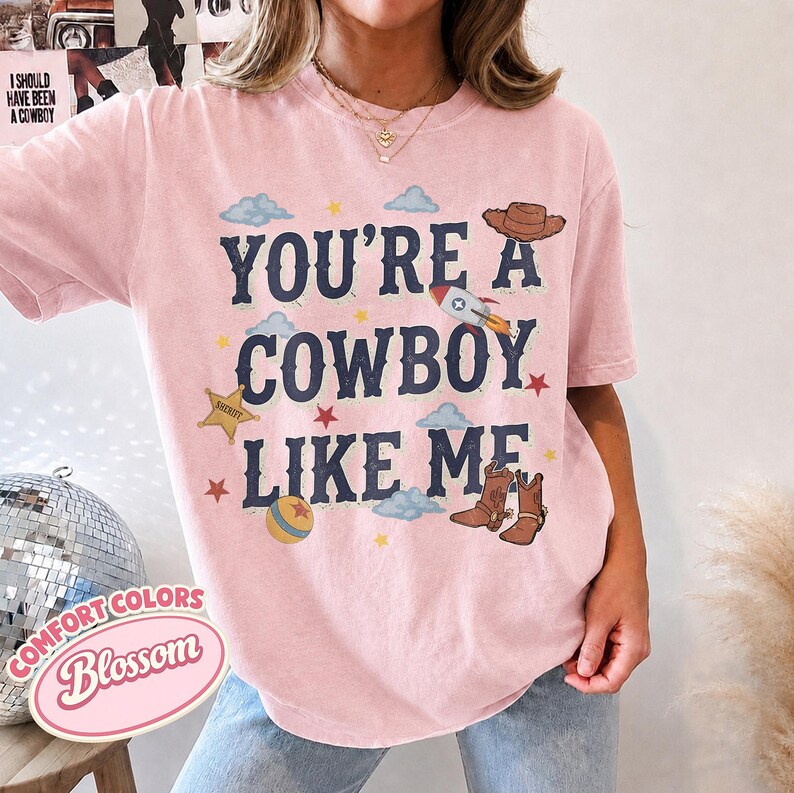 May include: A light pink short-sleeve t-shirt featuring the dark blue text YOU'RE A COWBOY LIKE ME. The graphic includes illustrations of a brown cowboy hat, a rocket ship, a gold sheriff badge, a ball, and brown cowboy boots with clouds and stars.