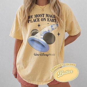 K&ouml;nnte beinhalten: Hellgelbes T-Shirt mit dem Text "THE MOST MAGICAL PLACE ON EARTH" und einer Grafik von Mickey Mouses Zaubererhut und Kugeln. Das Shirt tr&auml;gt auch die Worte "Walt Disney World" und "COMFORT COLORS Butter".