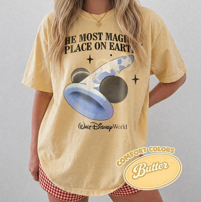 K&ouml;nnte beinhalten: Hellgelbes T-Shirt mit dem Text "THE MOST MAGICAL PLACE ON EARTH" und einer Grafik von Mickey Mouses Zaubererhut und Kugeln. Das Shirt tr&auml;gt auch die Worte "Walt Disney World" und "COMFORT COLORS Butter".