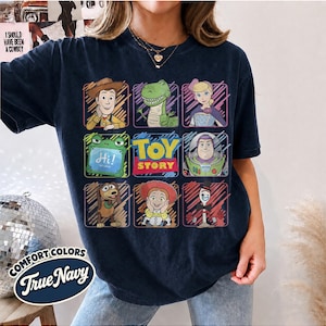 May include: A dark navy blue t-shirt featuring a grid of Toy Story characters including Woody, Rex, Bo Peep, Slinky Dog, Jessie, Forky, and Buzz Lightyear, with the Toy Story logo in the center and a Comfort Colors True Navy label graphic.