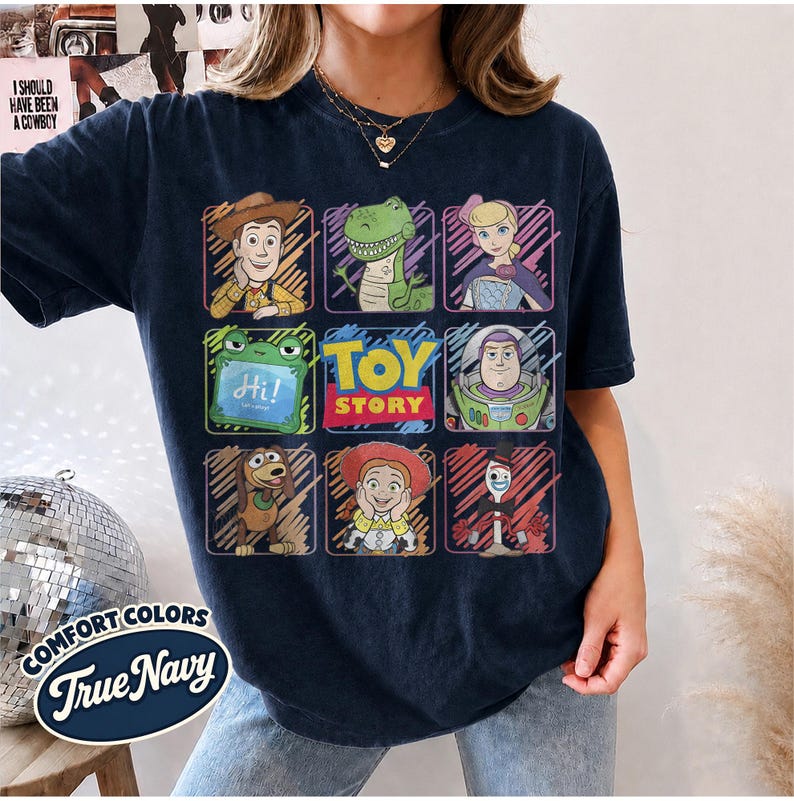May include: A dark navy blue t-shirt featuring a grid of Toy Story characters including Woody, Rex, Bo Peep, Slinky Dog, Jessie, Forky, and Buzz Lightyear, with the Toy Story logo in the center and a Comfort Colors True Navy label graphic.