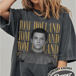 Peut inclure: T-shirt gris fonc&eacute; surdimensionn&eacute; avec l'impression dor&eacute;e "TOM HOLLAND" et un portrait. Le t-shirt a un aspect vintage et la marque "Comfort Colors Pepper" est imprim&eacute;e en bas &agrave; droite.