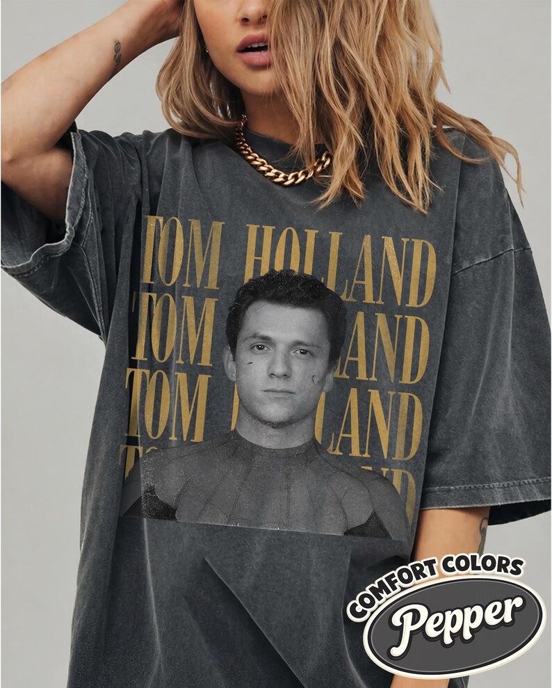 Peut inclure: T-shirt gris fonc&eacute; surdimensionn&eacute; avec l'impression dor&eacute;e "TOM HOLLAND" et un portrait. Le t-shirt a un aspect vintage et la marque "Comfort Colors Pepper" est imprim&eacute;e en bas &agrave; droite.