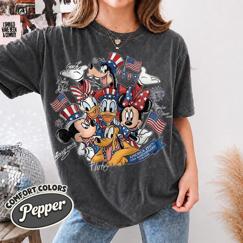 May include: Dark gray Comfort Colors t-shirt featuring a graphic of Disney characters Mickey Mouse, Minnie Mouse, Donald Duck, Daisy Duck, Goofy, and Pluto, celebrating the USA's 250th anniversary. The design includes American flags and patriotic colors.