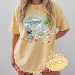 Op de afbeelding: Geel Comfort Colors T-shirt met een strandsc&egrave;ne-afbeelding. De afbeelding toont een cruiseschip, palmboom, zandkasteel, strandstoel en Mickey Mouse. Het shirt heeft de woorden "Comfort Colors" en "Butter".