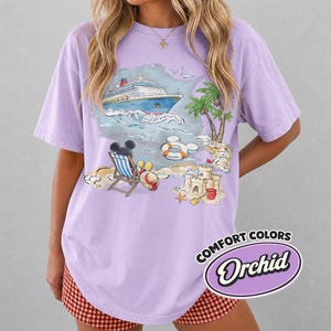 Op de afbeelding: Een lila Comfort Colors T-shirt met een strandsc&egrave;ne met een cruiseschip, palmbomen en een zandkasteel. Het shirt heeft een Mickey Mouse-afbeelding in een strandstoel. De tekst "COMFORT COLORS Orchid" staat rechtsonder.