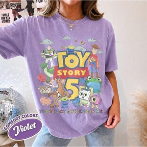 May include: A violet short-sleeve t-shirt featuring a graphic print of Toy Story characters including Buzz Lightyear, Woody, and Rex. The text reads Toy Story 5 and You've Got A Friend In Me. The shirt has a crew neckline and a relaxed fit design.