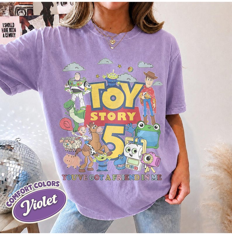 May include: A violet short-sleeve t-shirt featuring a graphic print of Toy Story characters including Buzz Lightyear, Woody, and Rex. The text reads Toy Story 5 and You've Got A Friend In Me. The shirt has a crew neckline and a relaxed fit design.