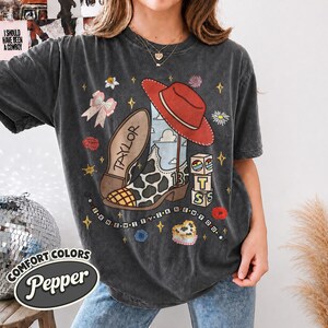 Peut inclure: Un t-shirt gris anthracite avec une impression graphique repr&eacute;sentant une botte de cow-boy &agrave; motifs de vache, un chapeau de cow-boy rouge, un n&oelig;ud rose et des blocs empil&eacute;s avec les lettres T, S et le chiffre 13. Le texte Taylor et I KNEW IT YIK NEW YOU est visible.