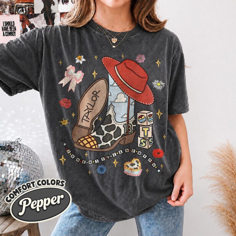 Peut inclure: Un t-shirt gris anthracite avec une impression graphique repr&eacute;sentant une botte de cow-boy &agrave; motifs de vache, un chapeau de cow-boy rouge, un n&oelig;ud rose et des blocs empil&eacute;s avec les lettres T, S et le chiffre 13. Le texte Taylor et I KNEW IT YIK NEW YOU est visible.
